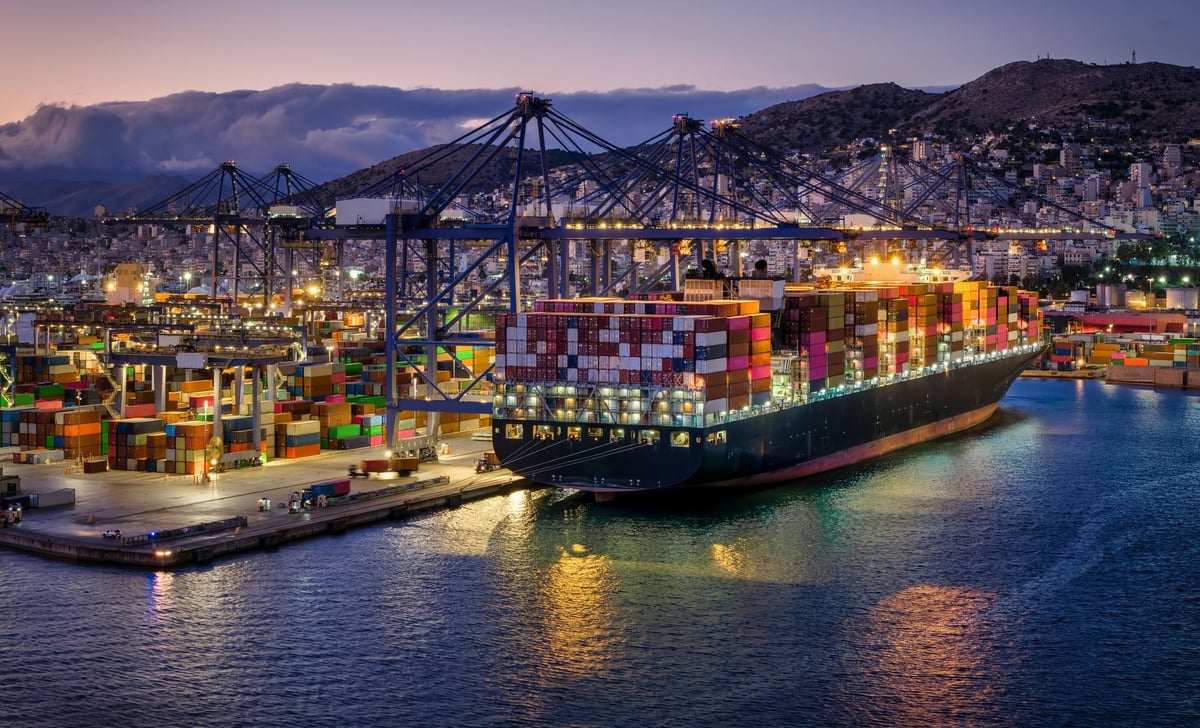 A illuminated, large container super cargo ship is being unloaded and loaded by cranes and trucks in a commercial terminal during evening